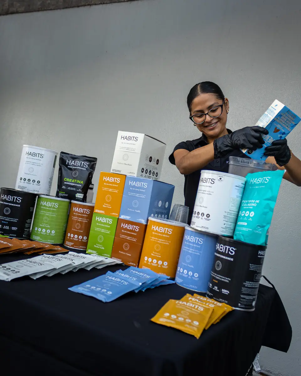 Una promotora de Habits Proteins muestra un sobre de producto sobre la mesa llena de botes y muestras.