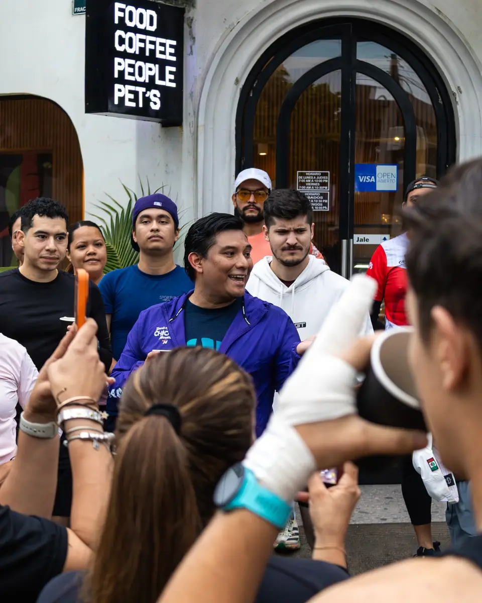 Corredores brindando con las manos en alto frente a la fachada de un café, con el letrero de «Food Coffee People Pets» detrás.