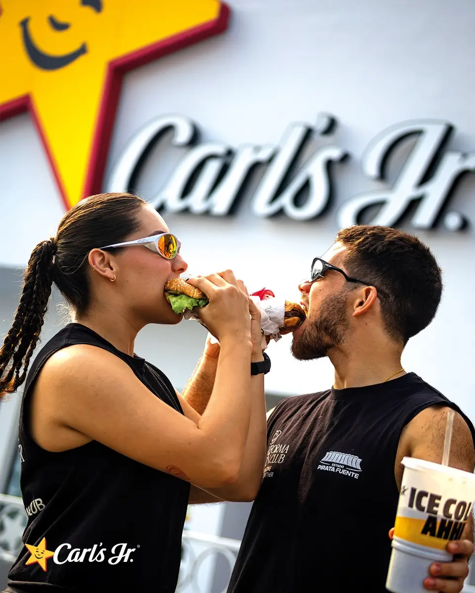 Una corredora y un corredor comparten un momento cómico comiendo frente al letrero de Carl's Jr.